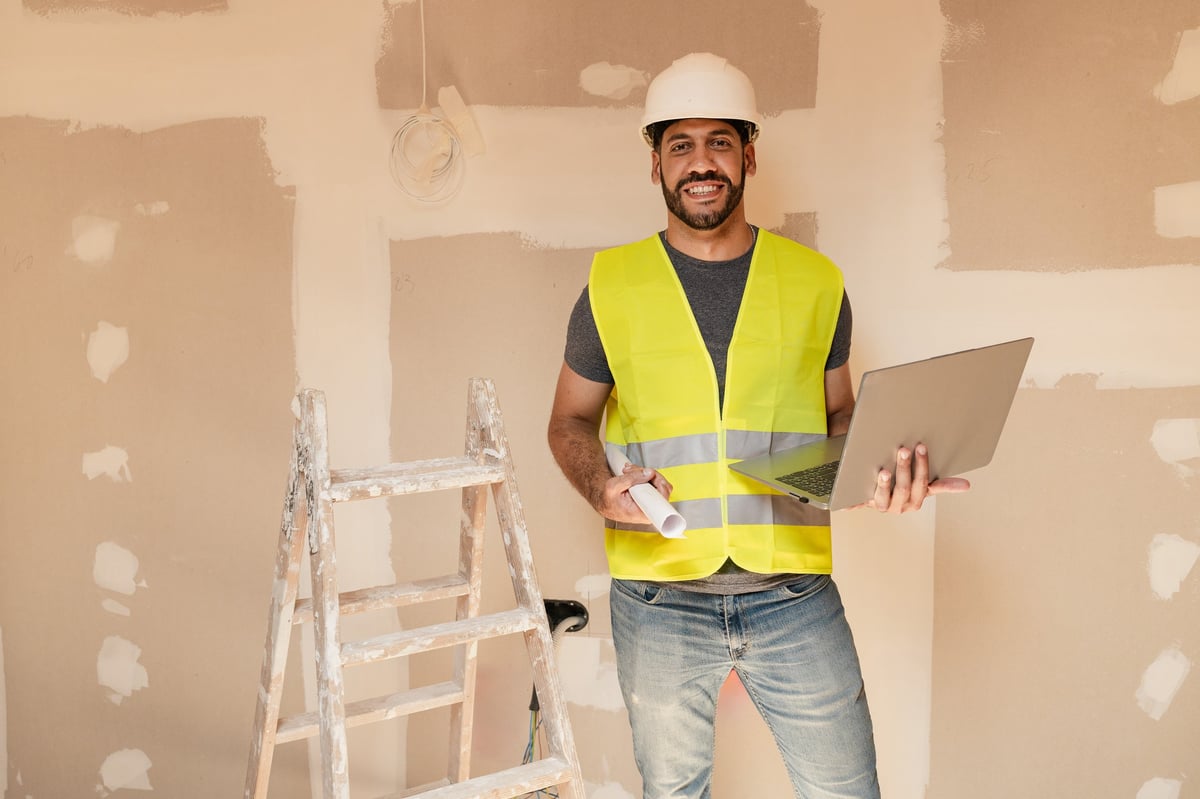 Hispanic male construction worker holding laptop and blueprint smiling at camera at building site