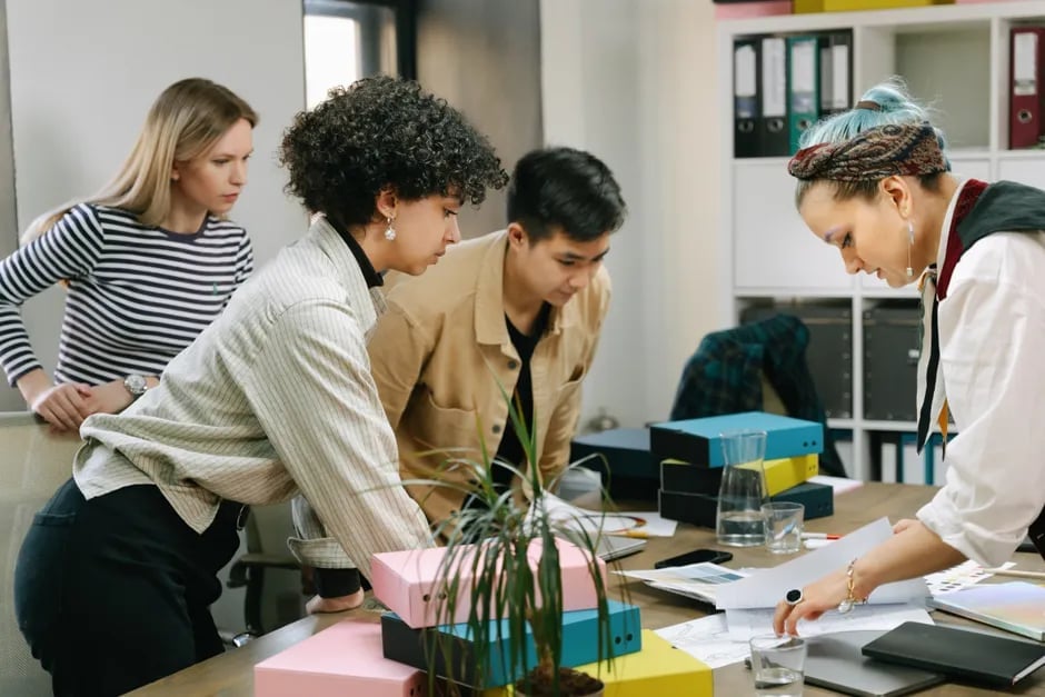 A diverse team of five professionals collaborating at a table in a modern office, reviewing documents and a potted plant.