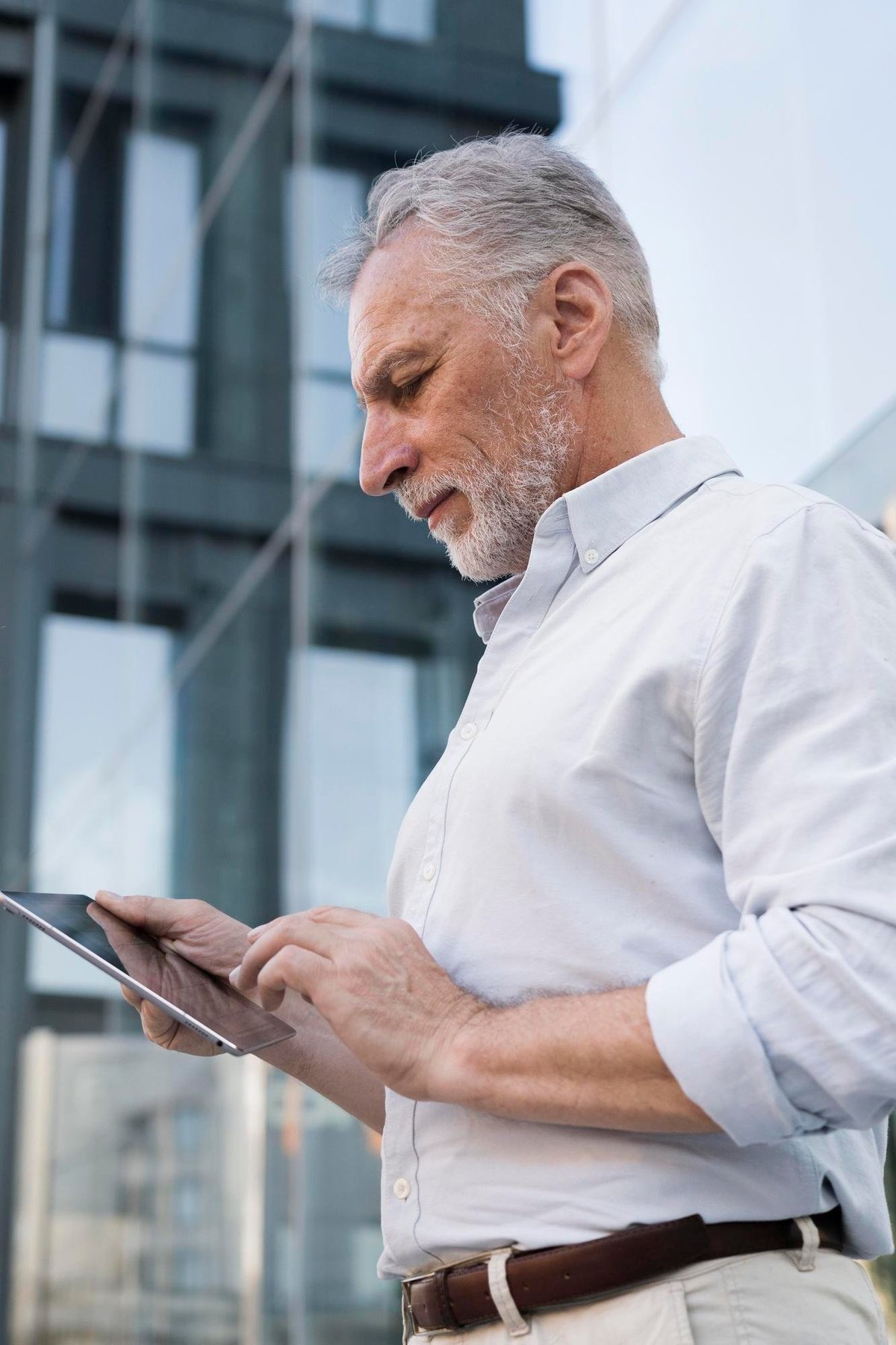 Mature businessman in white shirt using tablet outdoors with modern buildings in background