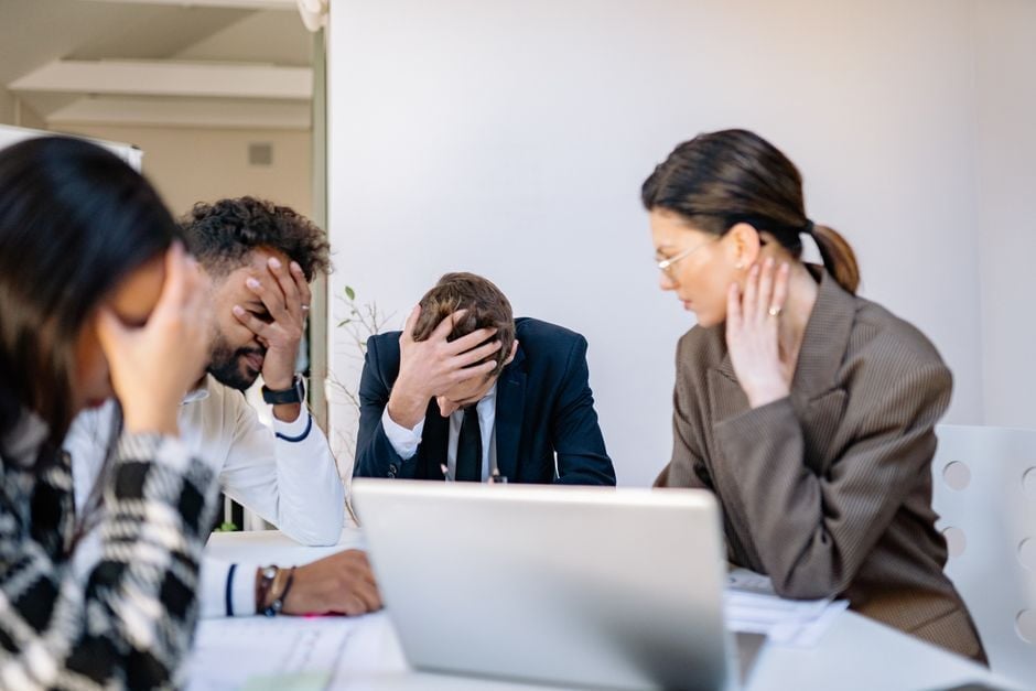 Four business professionals sitting at a table with a laptop, appearing stressed or discussing work matters in an office setting.